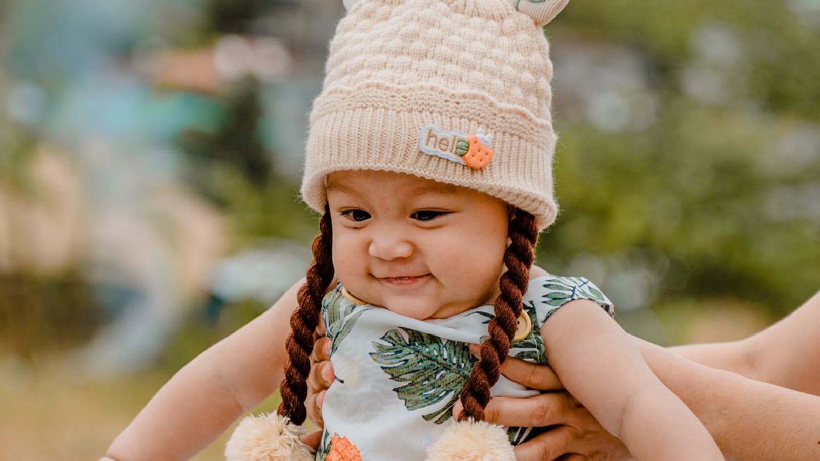A baby in a light romper standing with a parent's hands nearby