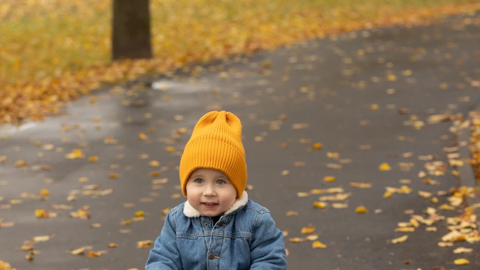 A toddler in a hat riding a yellow toy car along a leaf-covered path