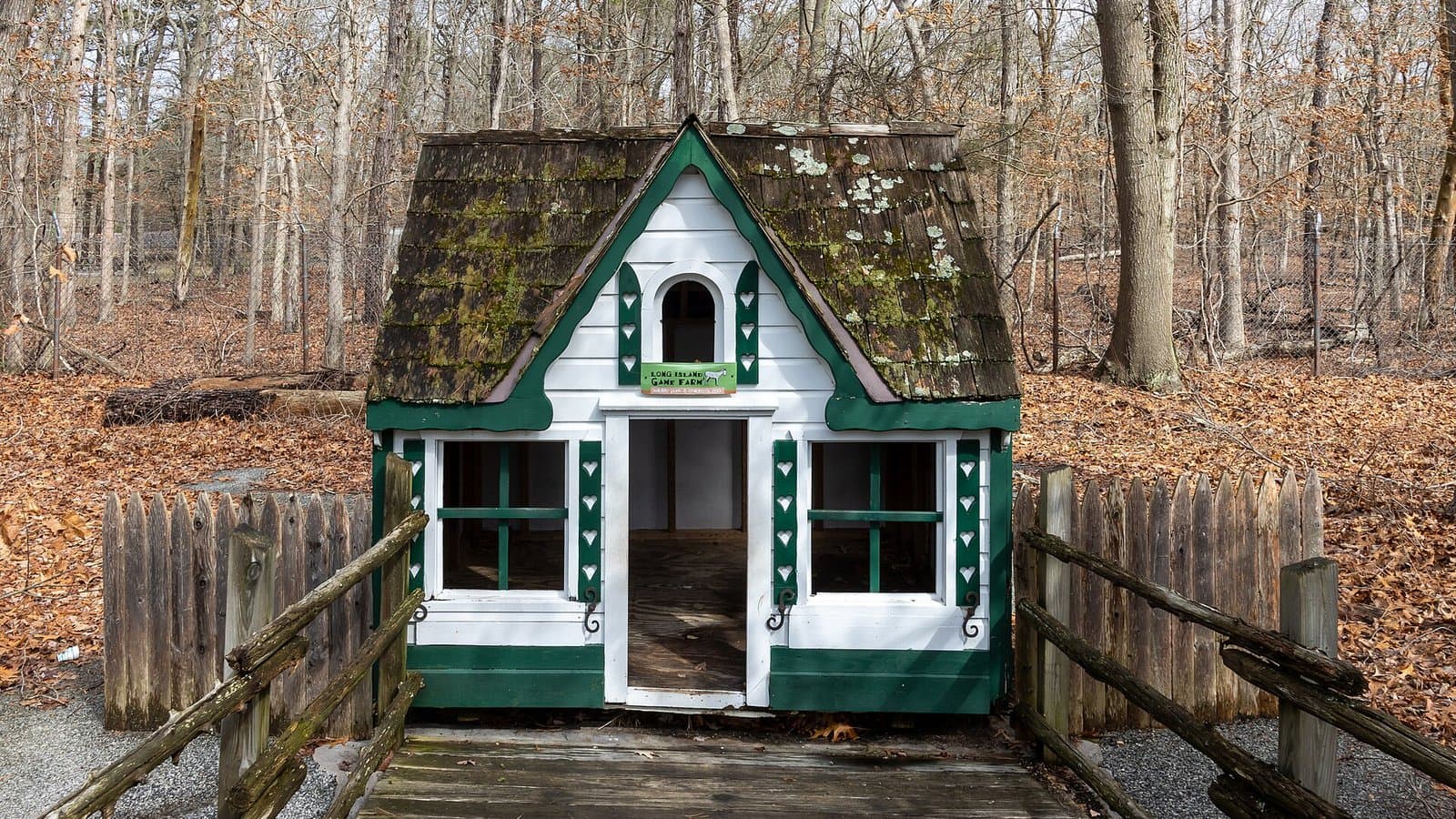 A white and green wooden outdoor playhouse at the end of a leaf-covered boardwalk in a wooded yard (photo: Mike Peel / Wikimedia Commons, CC BY-SA 4.0)