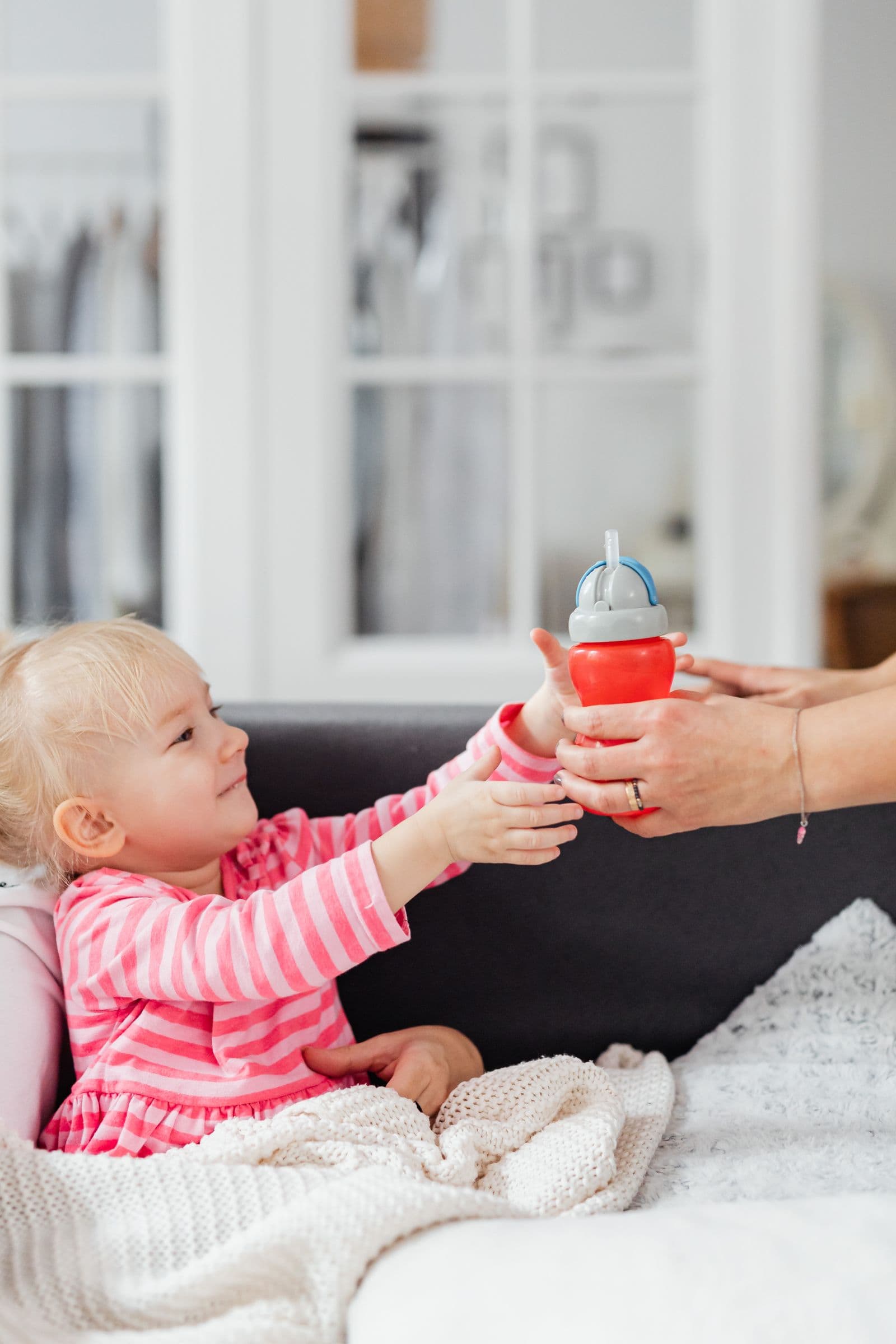 A smiling toddler in a pink striped top reaches for a straw sippy cup held by a parent