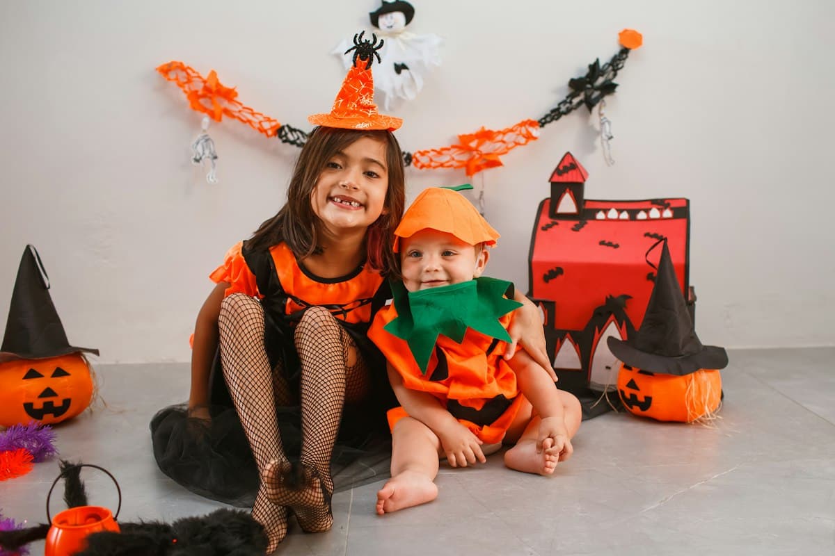 A smiling baby in a pumpkin costume sitting beside an older sister in a Halloween costume, with jack-o'-lanterns nearby