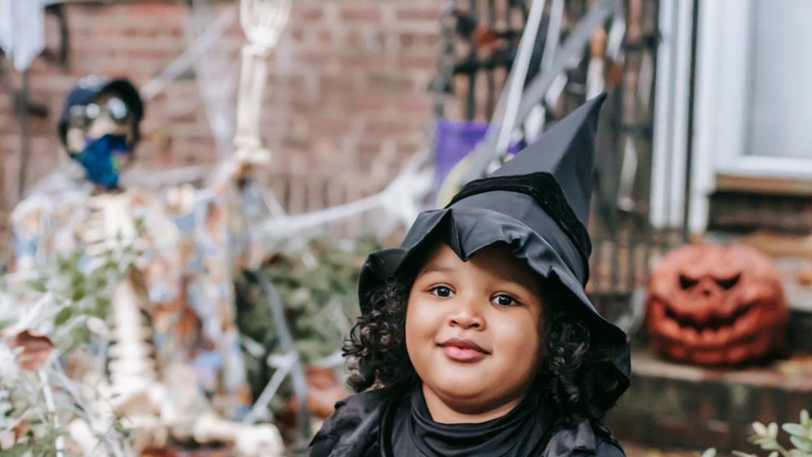 A young child in a black witch hat holding a pumpkin trick-or-treat bucket