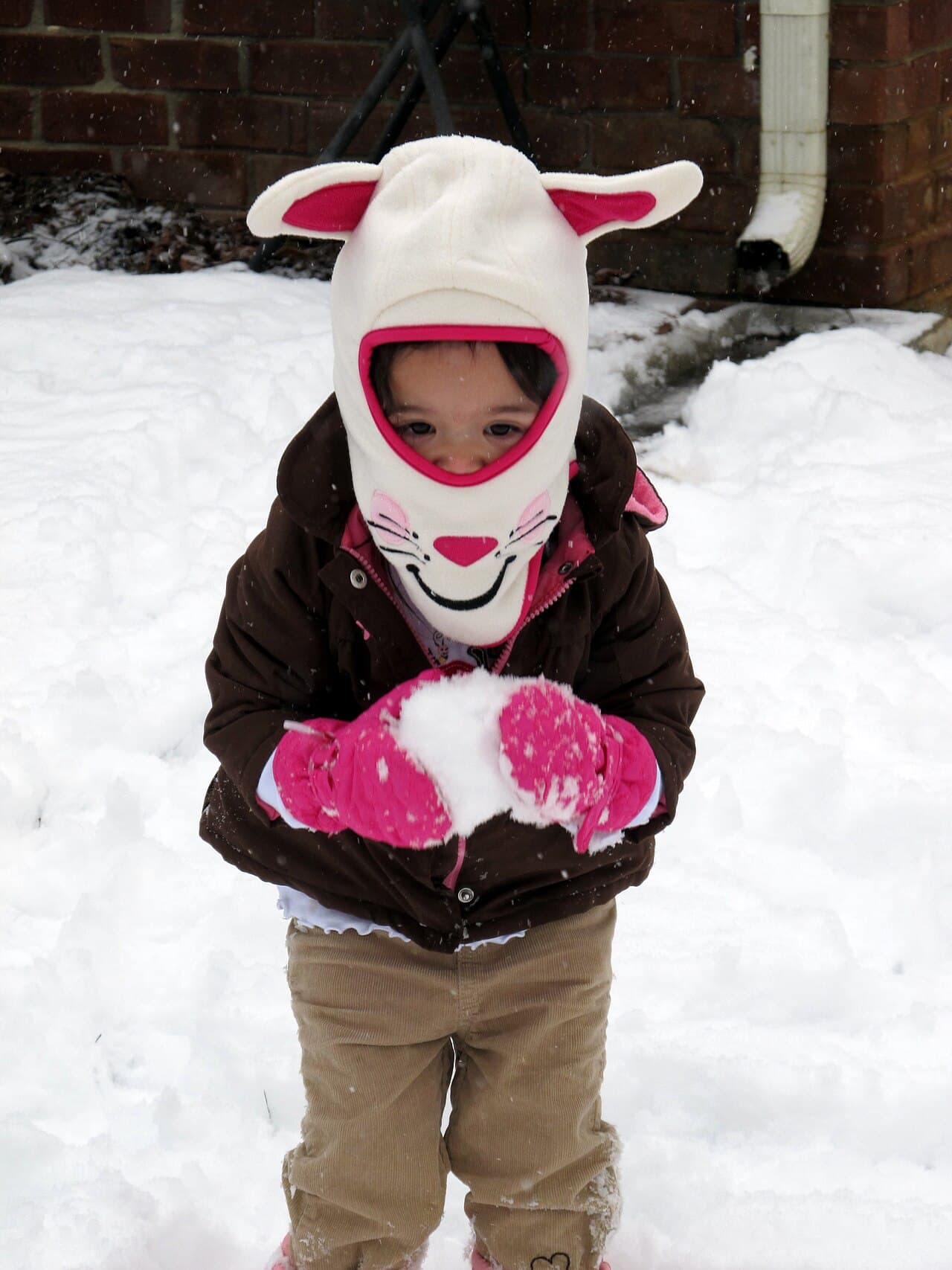 A toddler in a bunny balaclava and pink waterproof mittens packing a snowball in a snowy yard