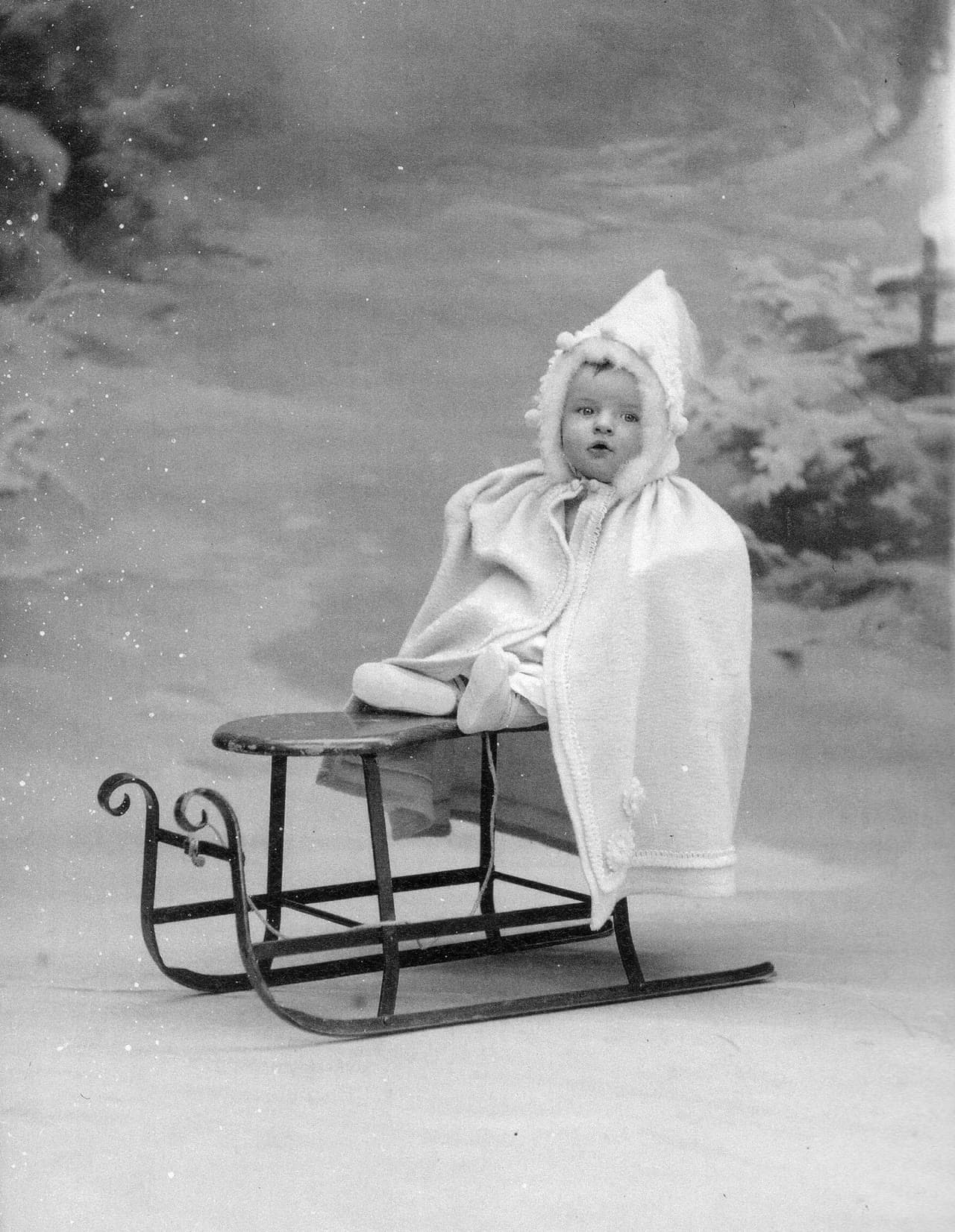 A toddler in a white hooded winter cape sitting on a small metal-runner sled in a snowy studio scene, 1901