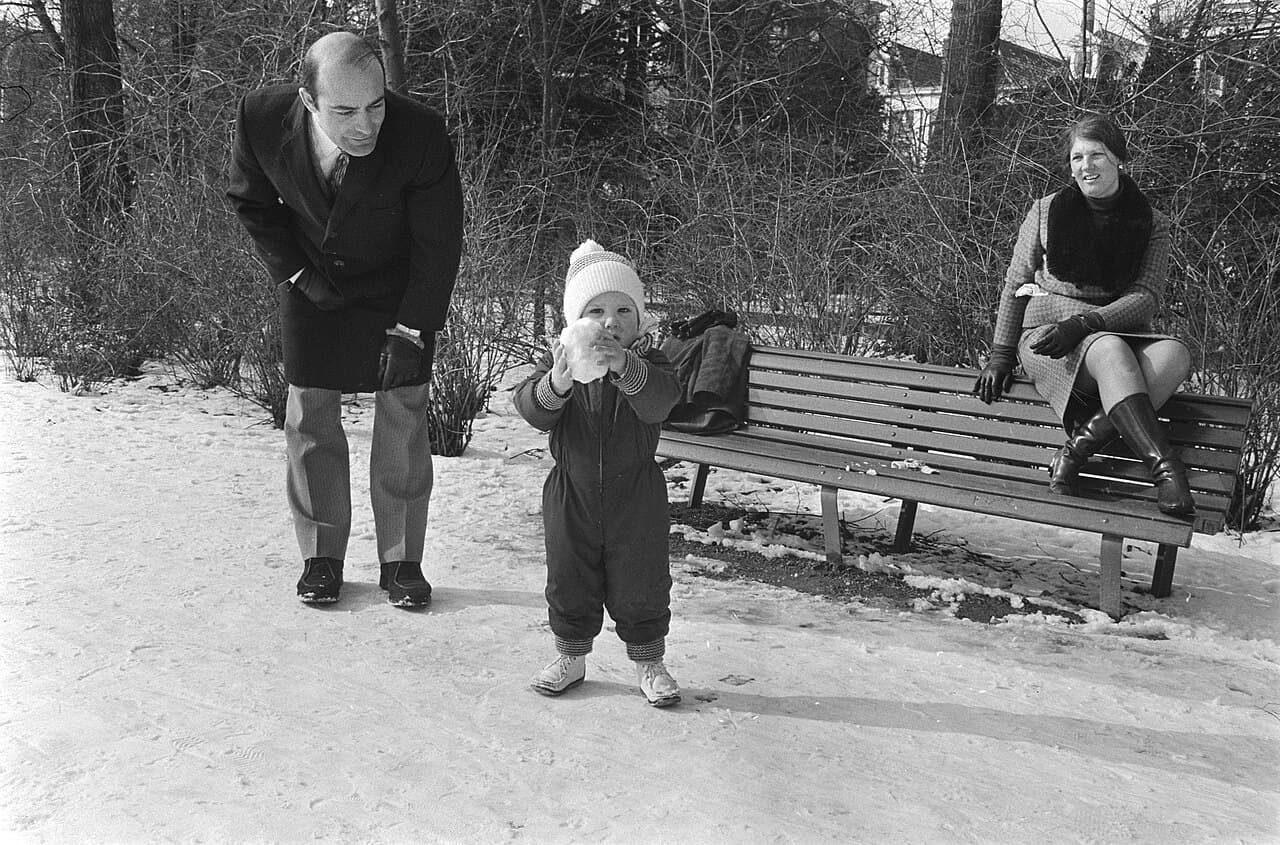 A toddler in a knit hat, snowsuit and winter boots standing in the snow in a park while two adults watch