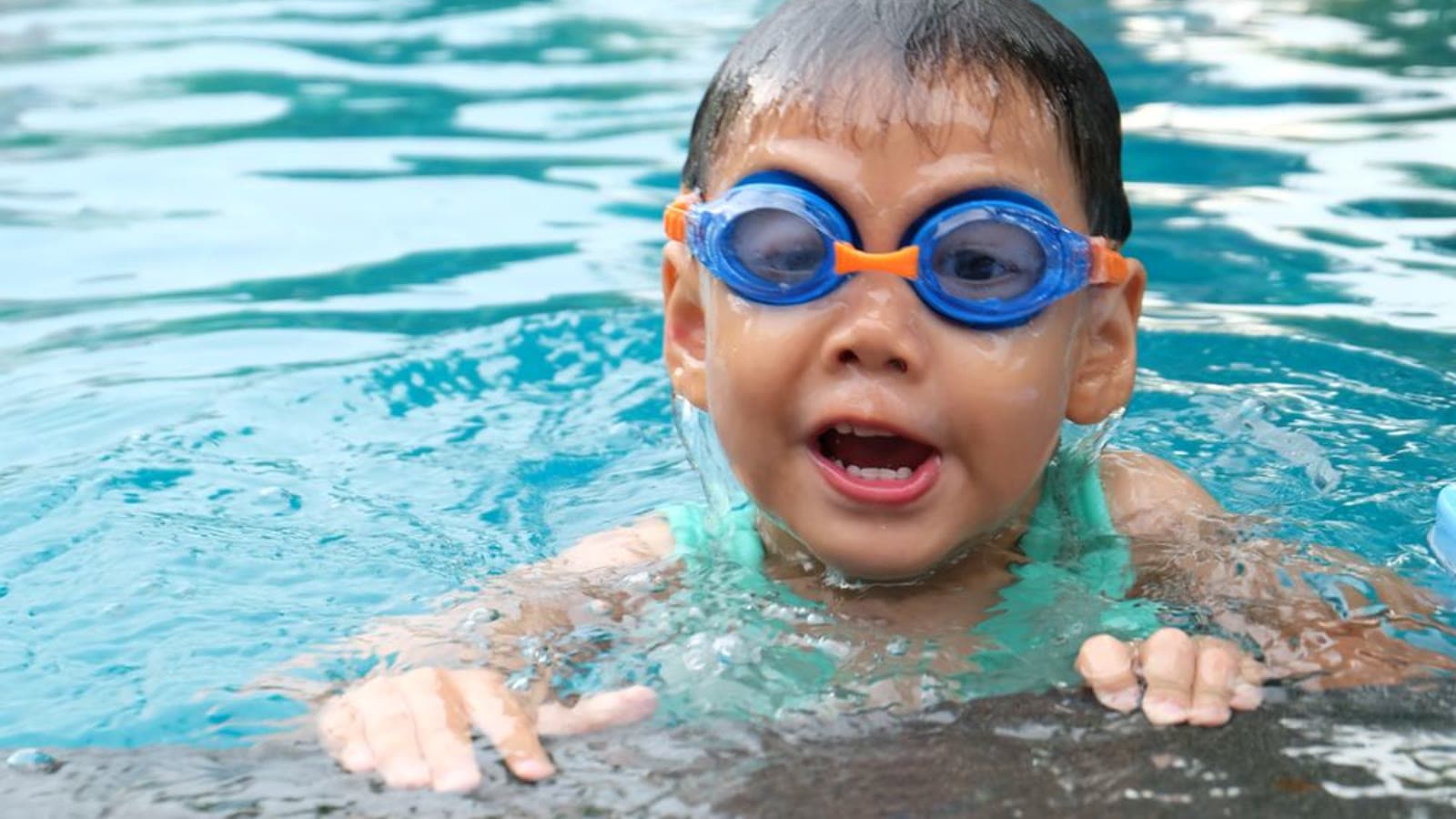 A toddler in a swimsuit and blue goggles swimming in a pool
