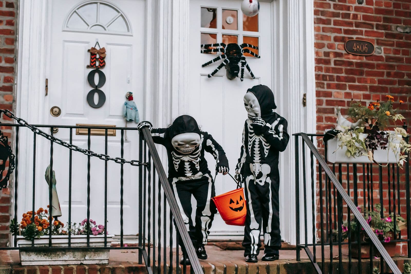 Two children in skeleton costumes walking down front steps on Halloween, one carrying an orange jack-o'-lantern treat bucket