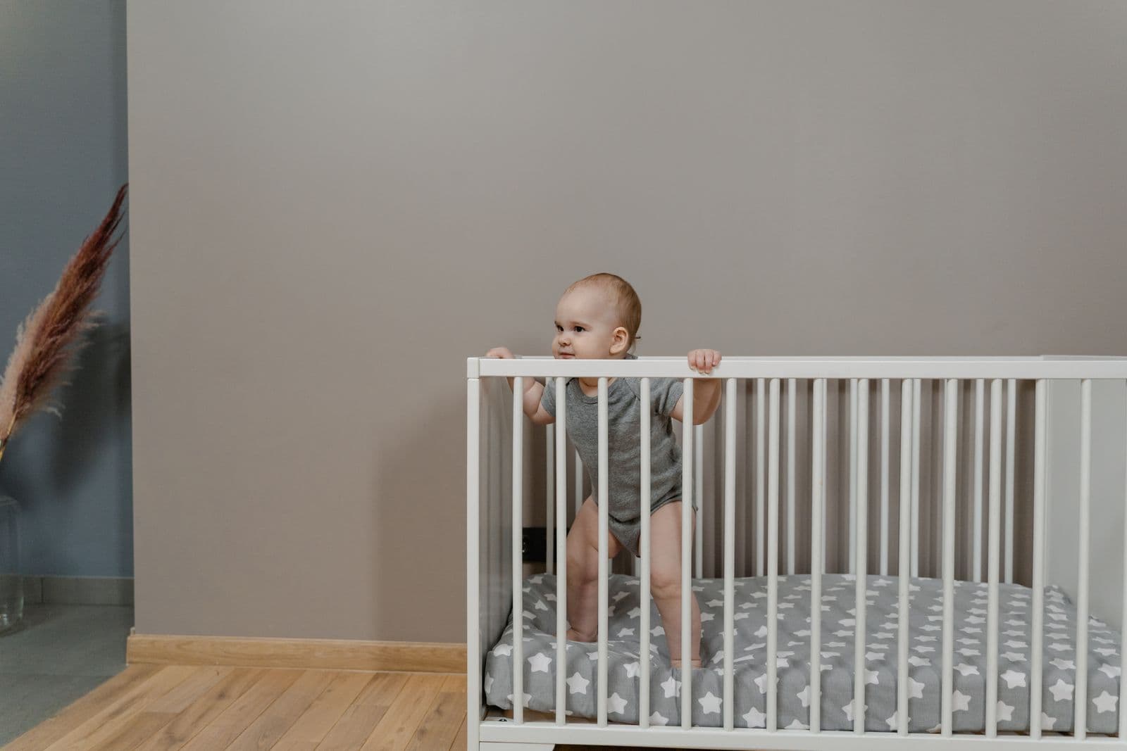 A baby standing and holding the rail of a crib in a bright room