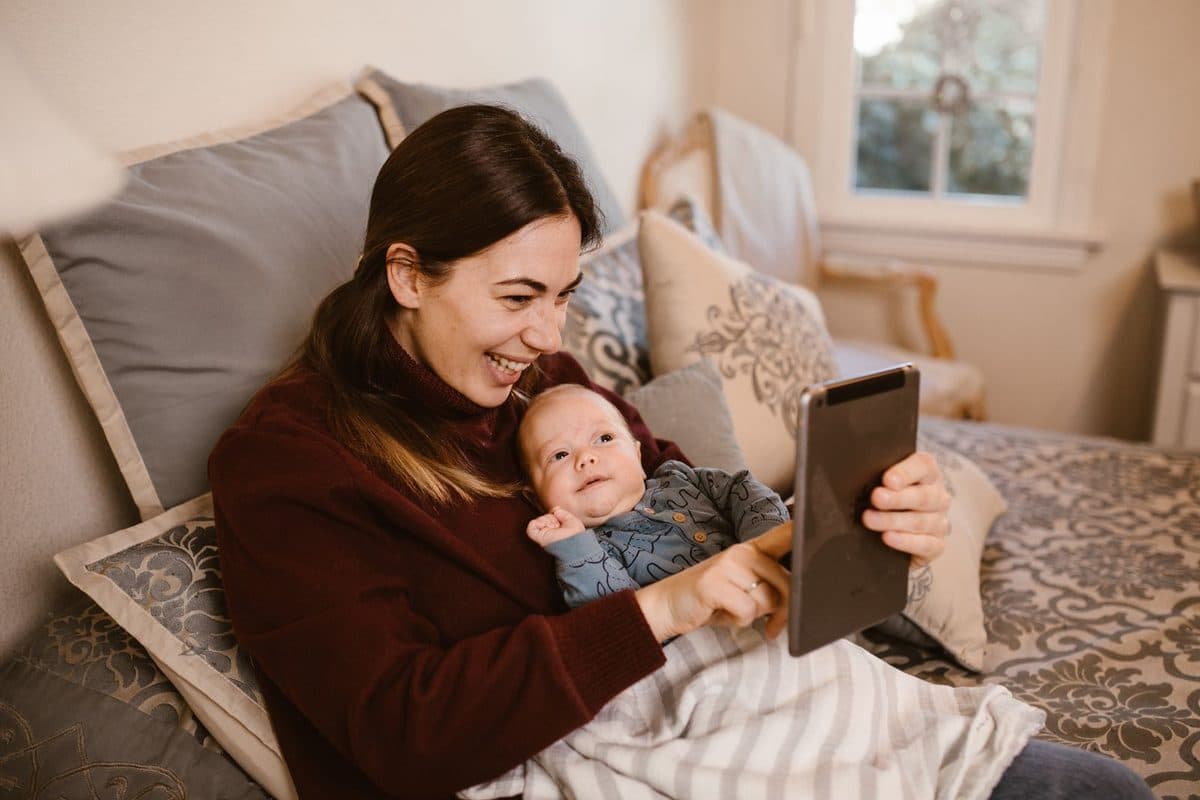 A smiling mother holds her baby on a bed in natural daylight while video calling family on a tablet